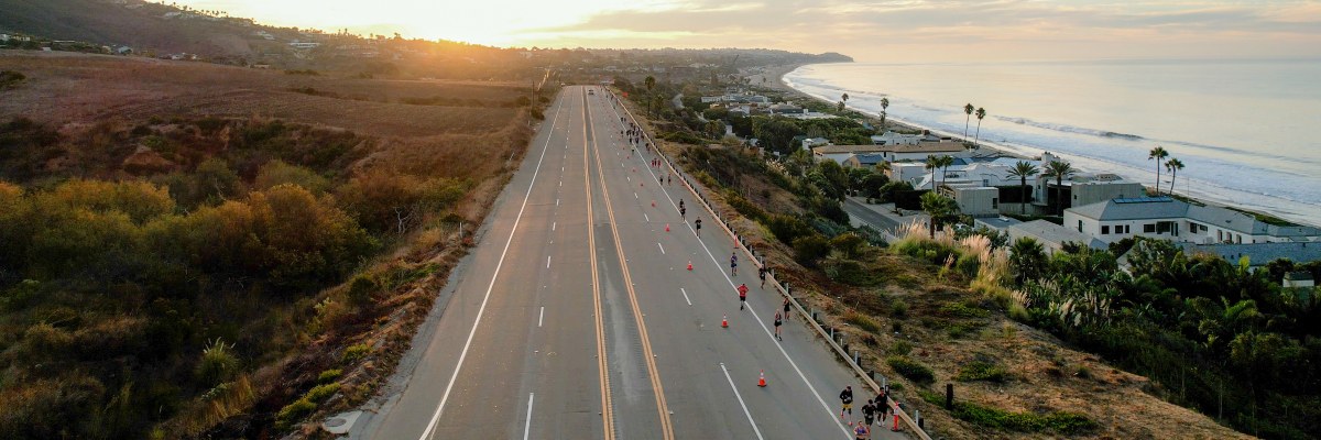 Aerial view of participants moving along a closed stretch of Pacific Coast Highway in Malibu at sunrise, with the Pacific Ocean on one side, coastal hills on the other, and golden morning light illuminating the coastline.